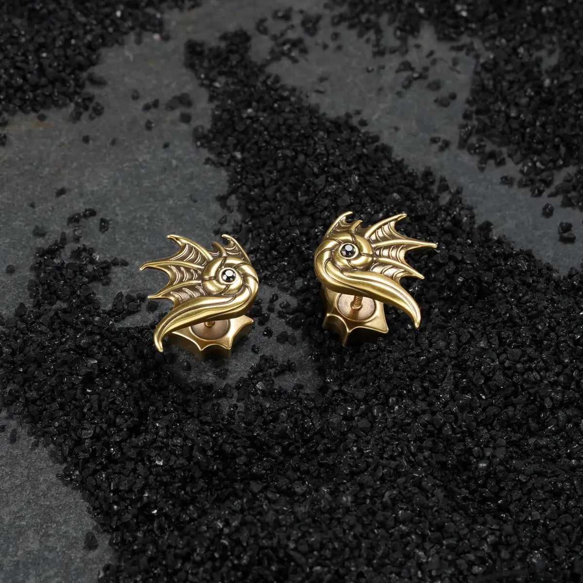 Gold Abyss Conch Ear Studs displayed on black sand, highlighting the curled conch shape, fin-like ridges, and black zircon detail.