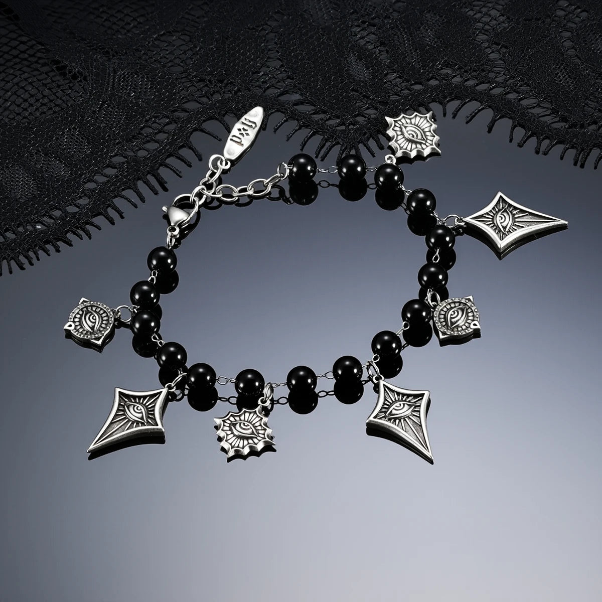 A gothic-style charm bracelet featuring black beads, silver-toned chain links, and spiderweb motifs, displayed on a dark reflective surface with a black lace background.