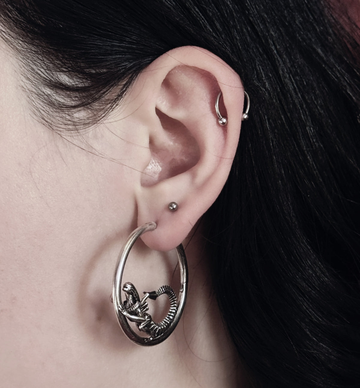 Close-up of a silver-tone hoop earring with alien skeleton design worn alongside a cartilage hoop and stud