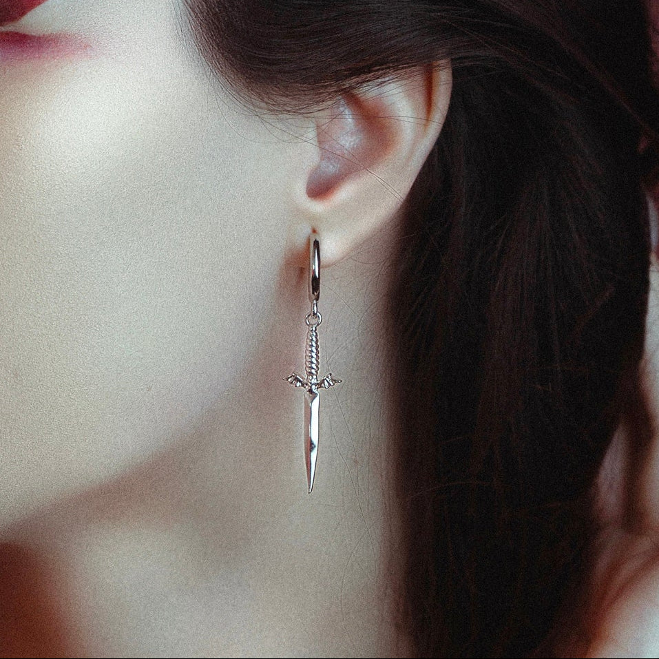Silver-tone dagger-shaped earring with winged hilt hanging from a hoop, worn on model's ear