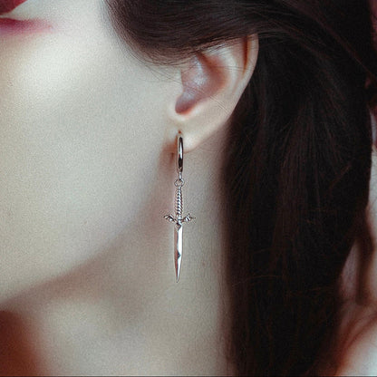 Silver-tone dagger-shaped earring with winged hilt hanging from a hoop, worn on model's ear