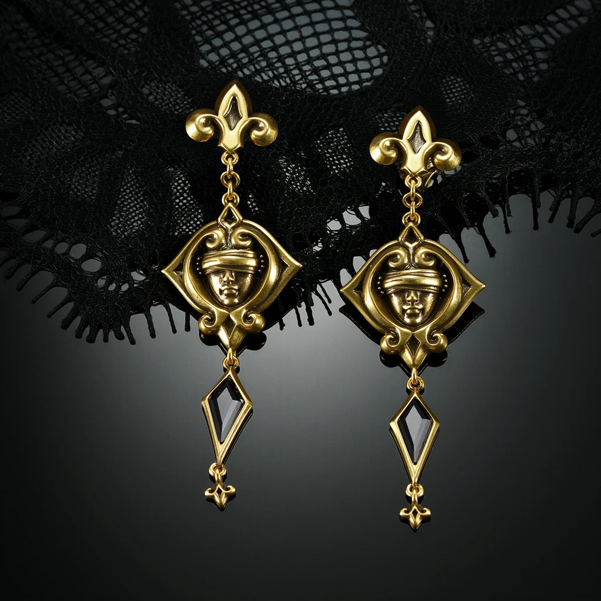 Gold-tone Blind Oracle Drop Earrings displayed upright against black lace on a dark reflective surface.