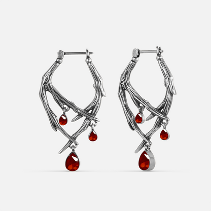 Silver-tone thorn-shaped hoop earrings pair with dangling red teardrop crystals, front view on white background