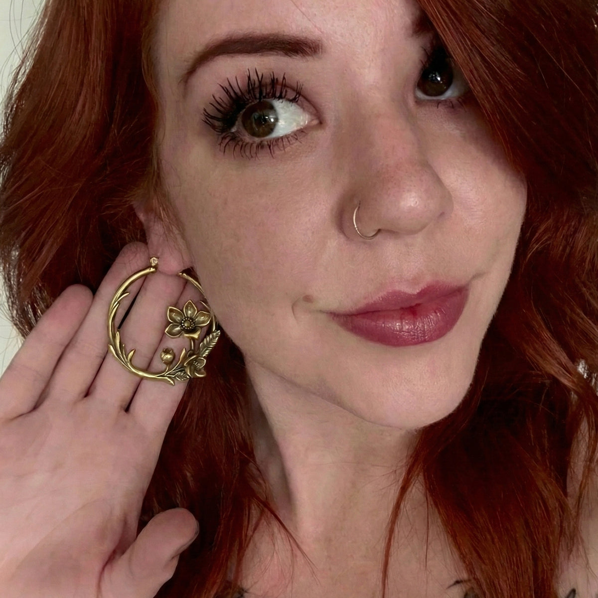Model with red hair holds up a gold-tone hoop earring with a floral vine design near the face