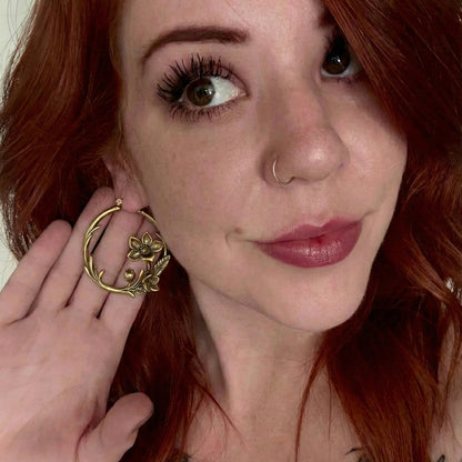 Model with red hair holds up a gold-tone hoop earring with a floral vine design near the face