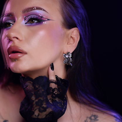 Close-up of a model wearing a silver-tone ginkgo leaf stud earring with a clear stone, styled with dramatic makeup