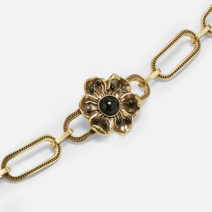 Close-up of a gold-tone flower charm with black stone accents on a paperclip chain bracelet