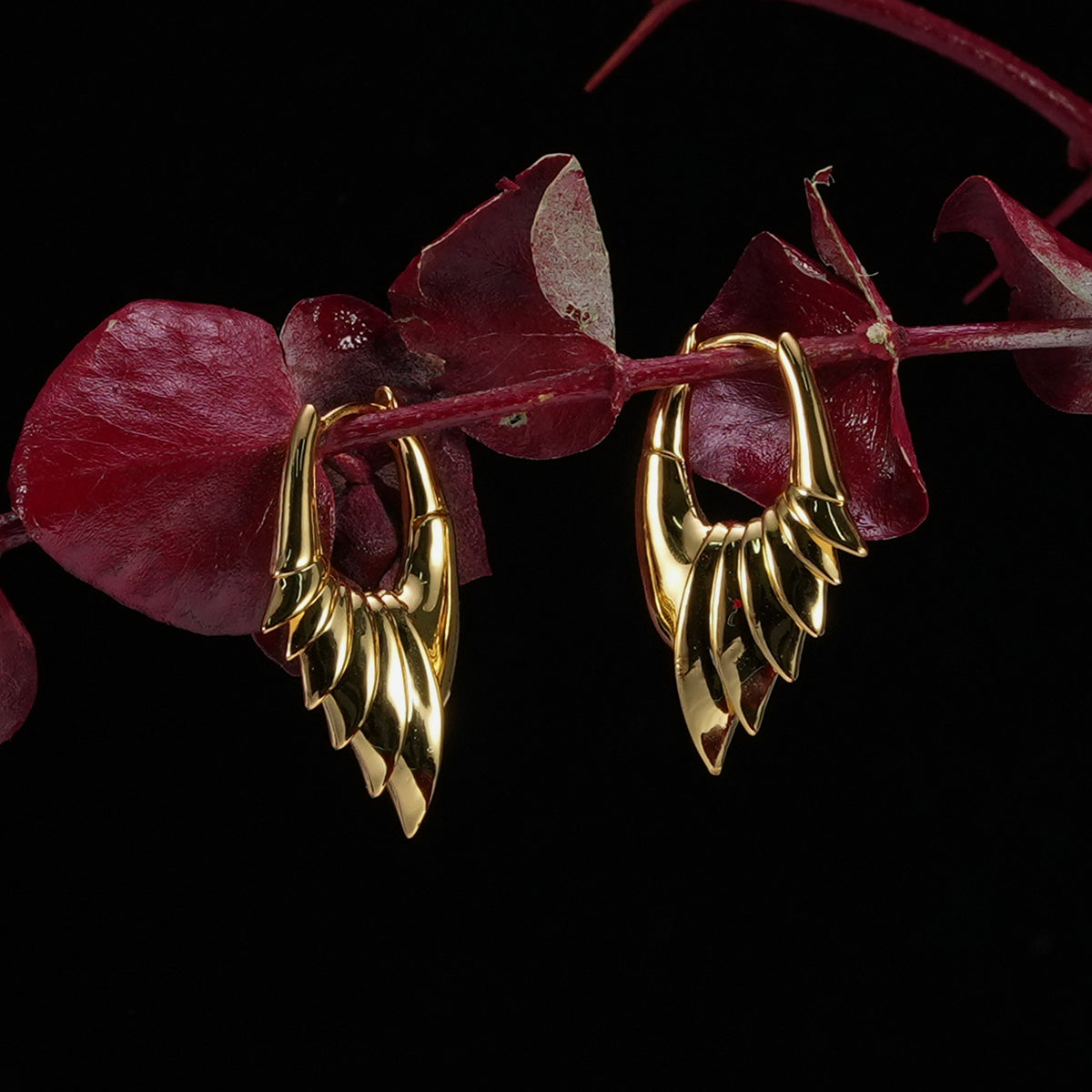 Pair of gold Carapace Wing hoop earrings hanging from red leaves