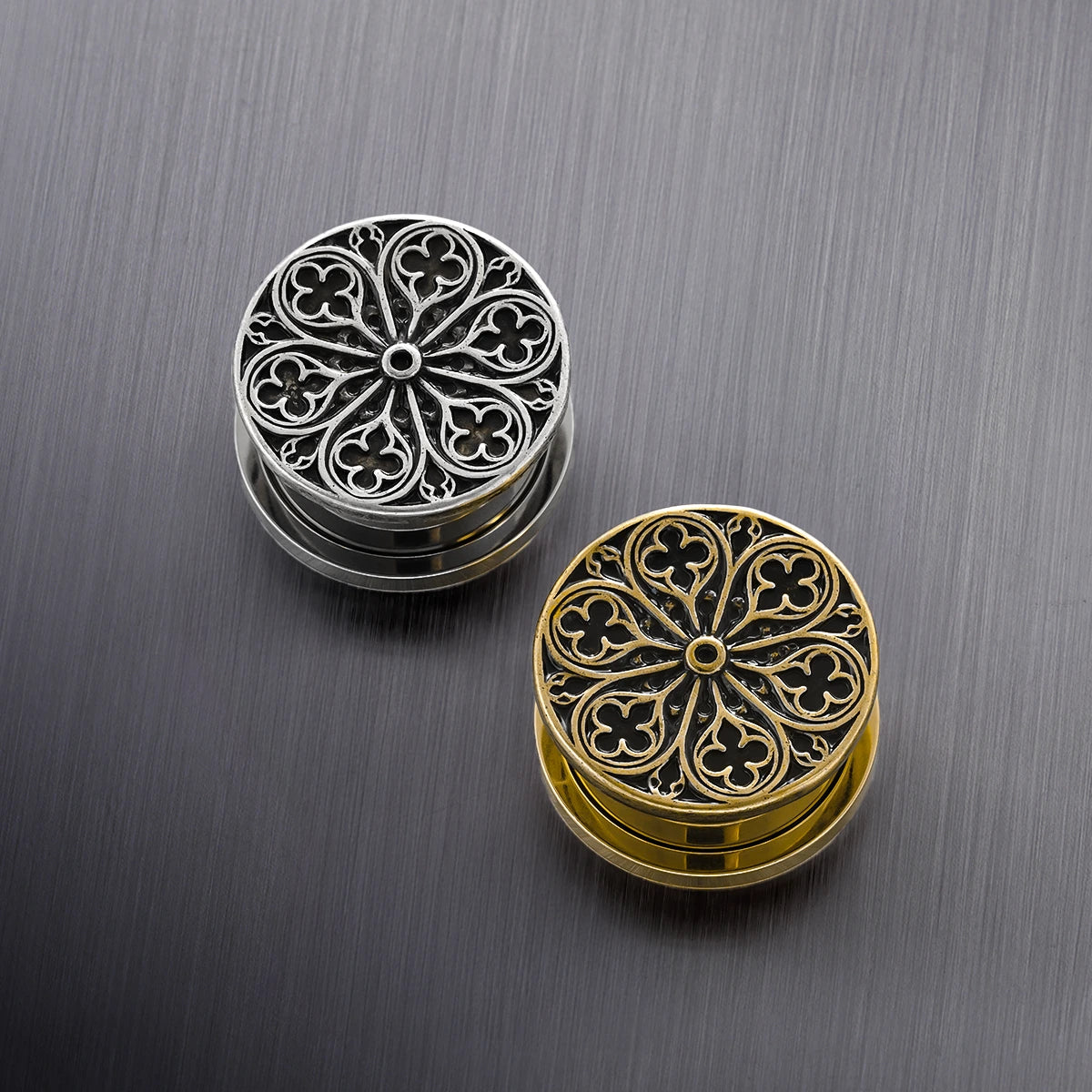 Silver-tone and gold-tone rose window ear plugs placed together on a brushed metal surface