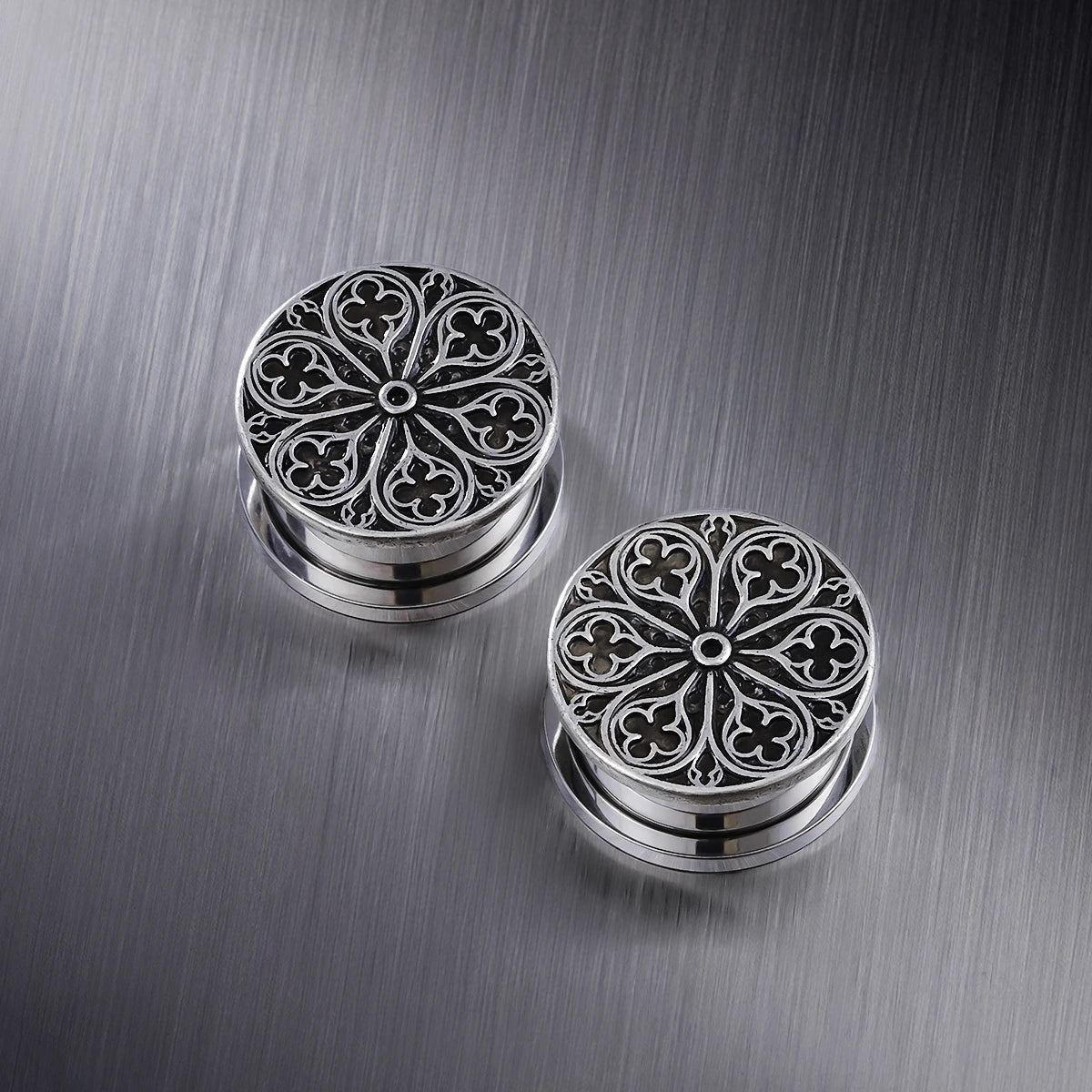 Pair of silver-tone ear plugs with a detailed cathedral rose window pattern on a metal surface