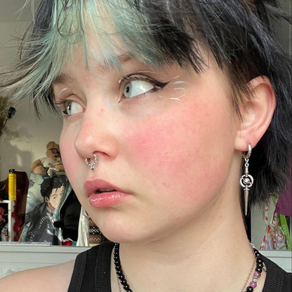 Celestial Orbit Earrings in 925 sterling silver, worn in a selfie with short green-gray hair, facial piercings, and a black b