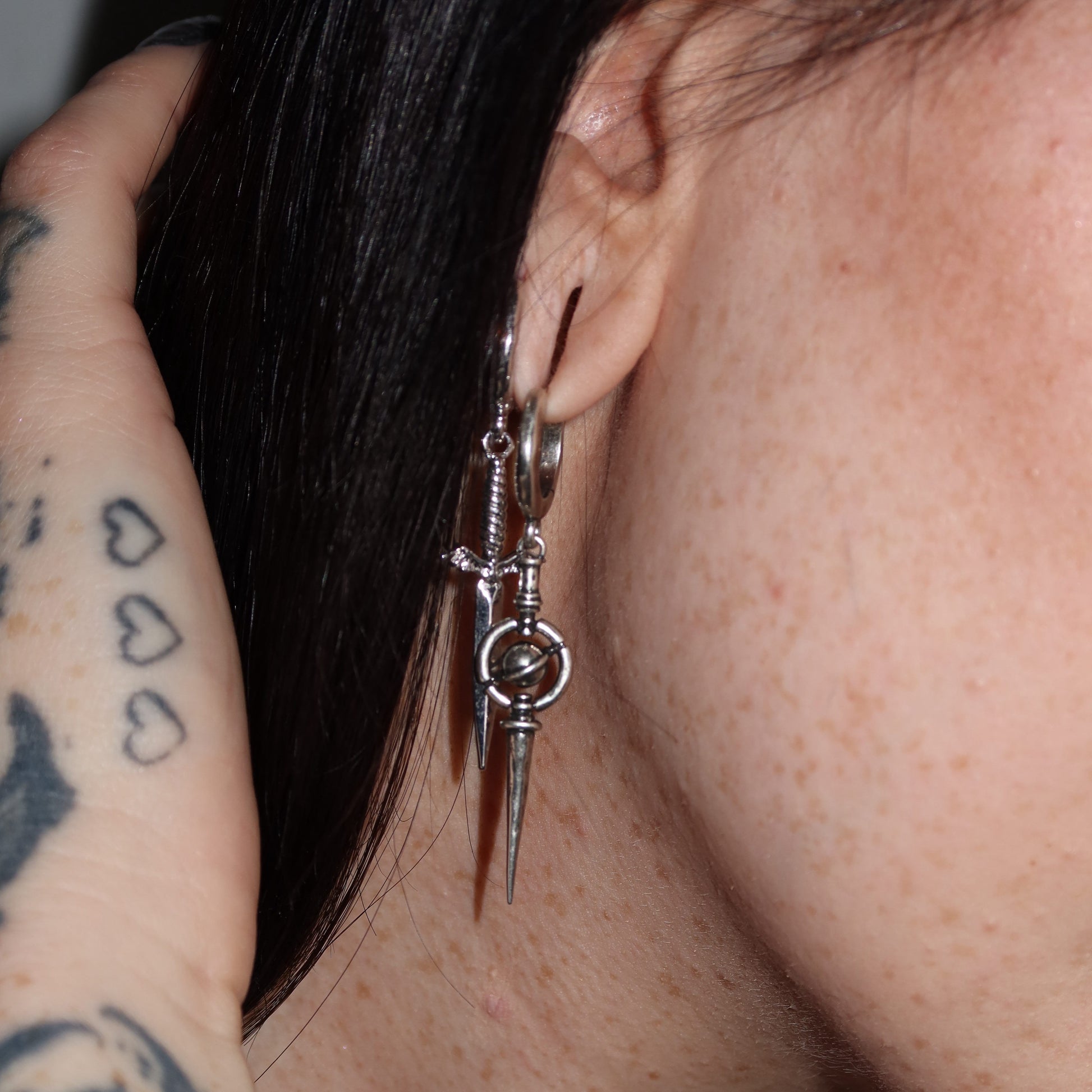 Silver-tone hoop earring with dagger and orbit ring charms worn on a model with dark hair, close side view