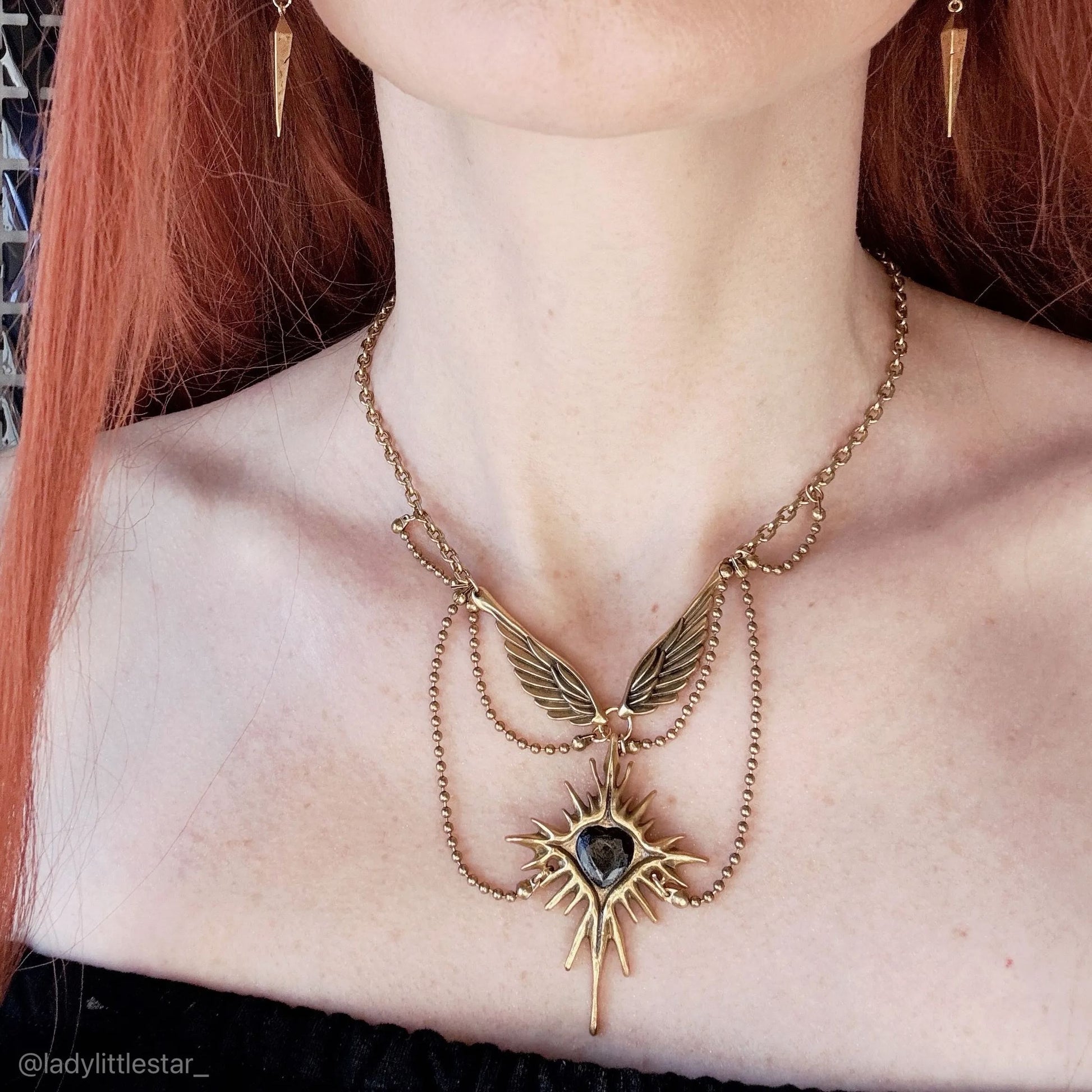 Gold-tone Celestial Starburst Wings necklace worn at the collarbone with layered chains and a black heart center