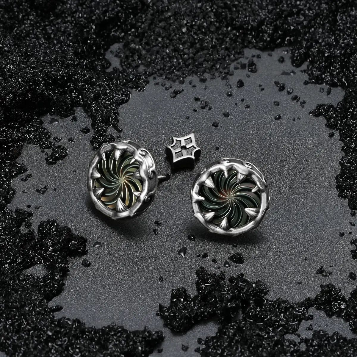 Silver Charybdis Maw Ear Studs displayed on black sand, highlighting the abalone shell spiral center and carved gothic rim details.