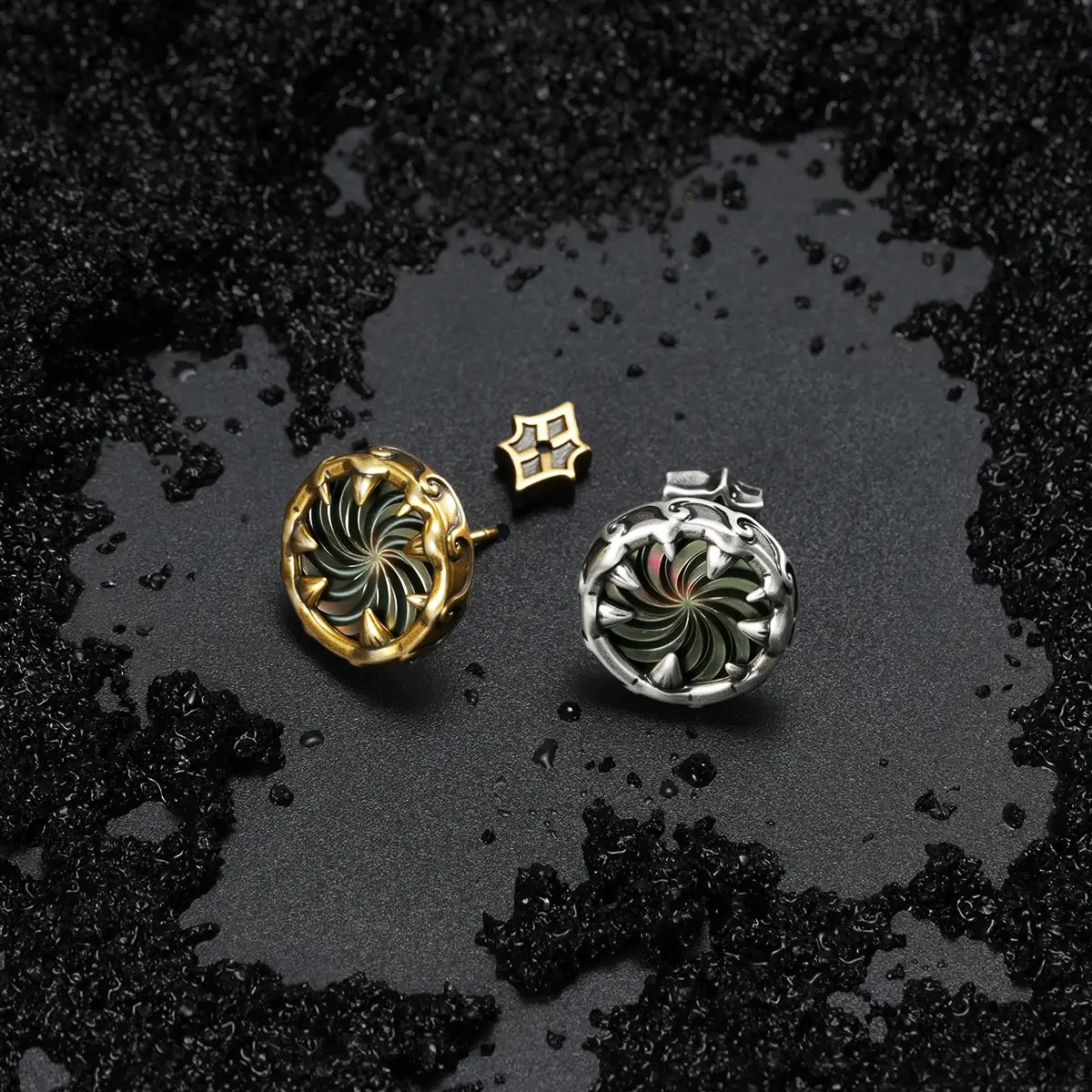 Silver and gold Charybdis Maw Ear Studs displayed on black sand, showing the swirling abalone shell centers and dark ocean-inspired styling.