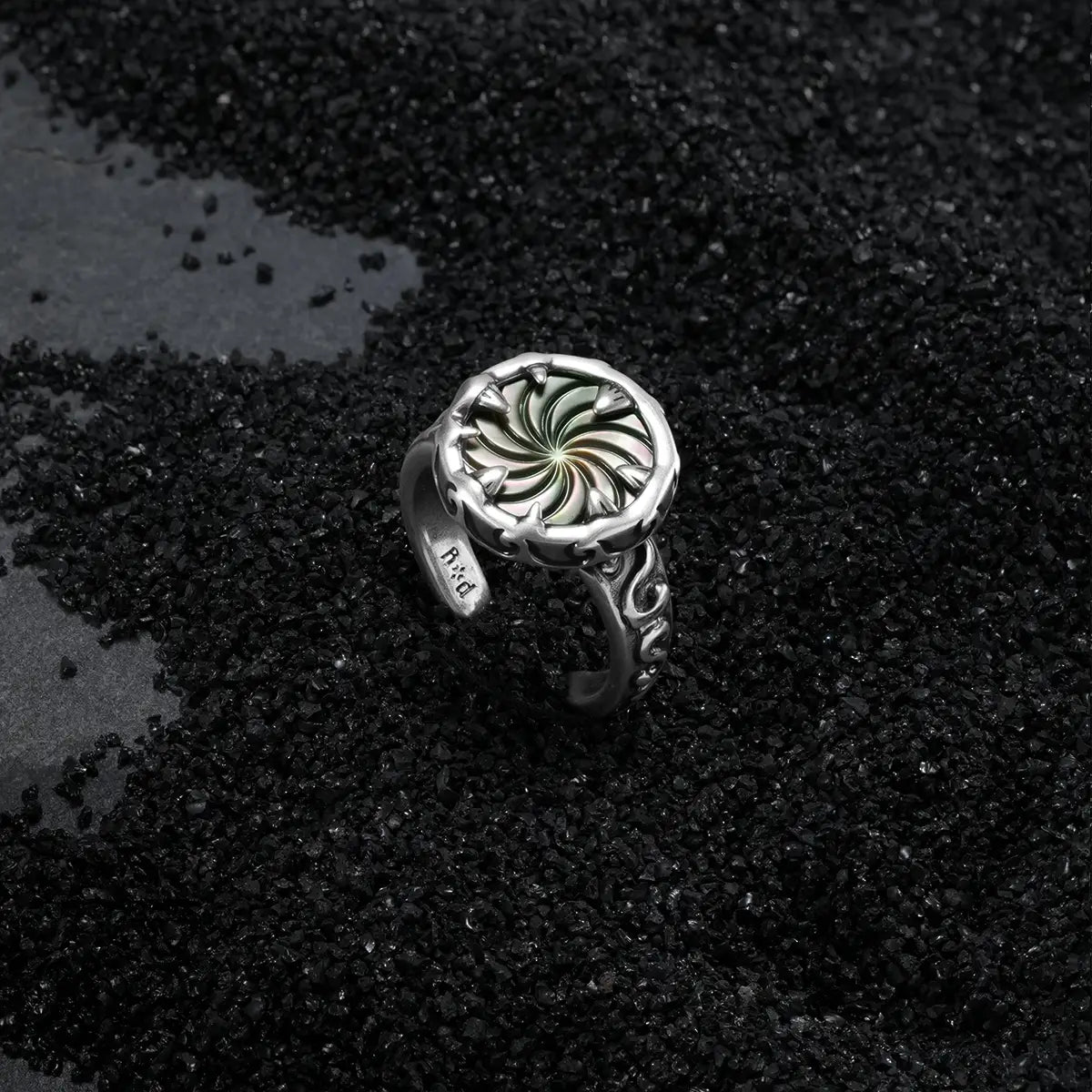 Charybdis Maw Ring displayed on black sand, featuring a swirling abalone shell center and carved antiqued silver details.