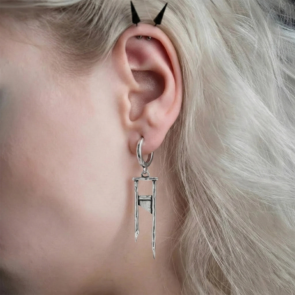 Silver-tone hoop earring with a guillotine-shaped dangling charm, worn on model's ear, close-up side view