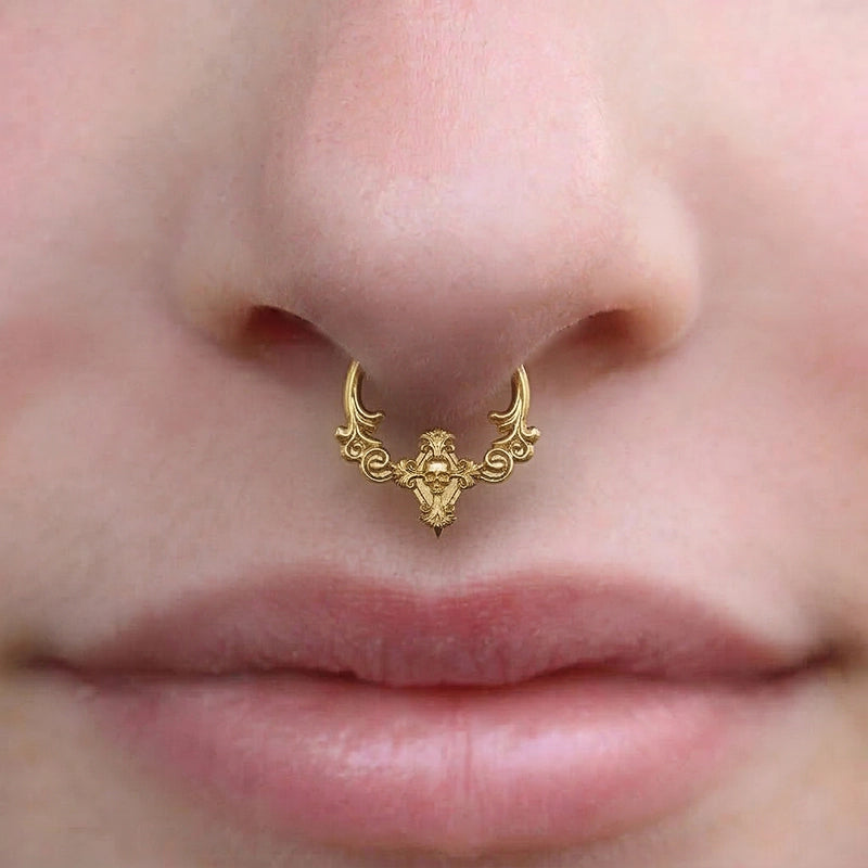 Gold-tone septum clicker with scrollwork and skull detail worn on a model's nose, close-up front view