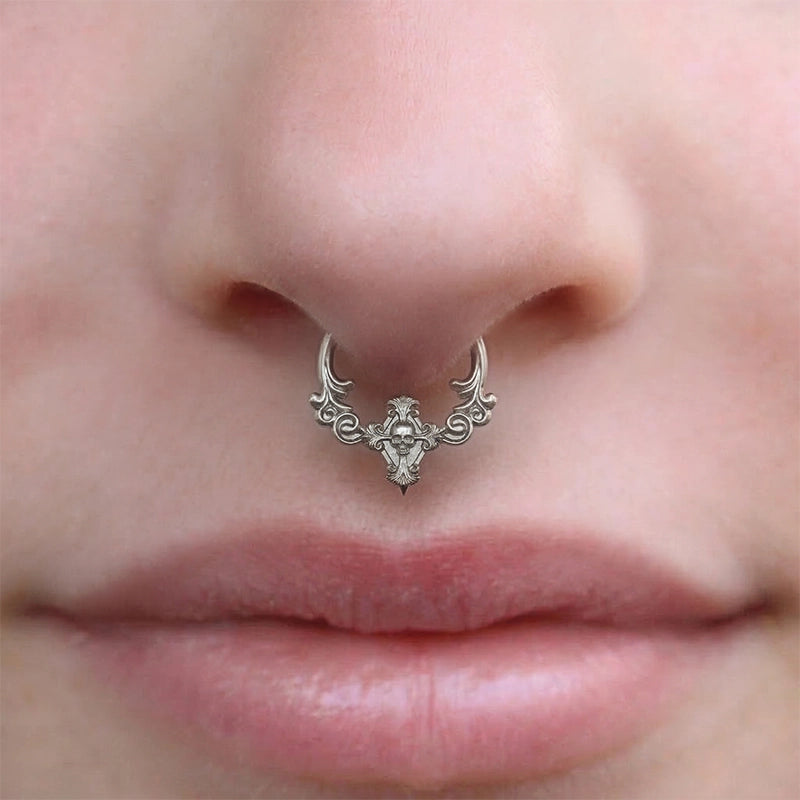 Silver-tone septum clicker with scrollwork and skull detail worn on a model's nose, close-up front view