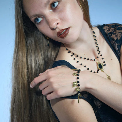 A close-up lifestyle shot of a model wearing a gothic-style jewelry set featuring black beaded strands with gold-toned charms, including a crescent moon, a star, and a green-embellished pendant, against a solid light blue background.