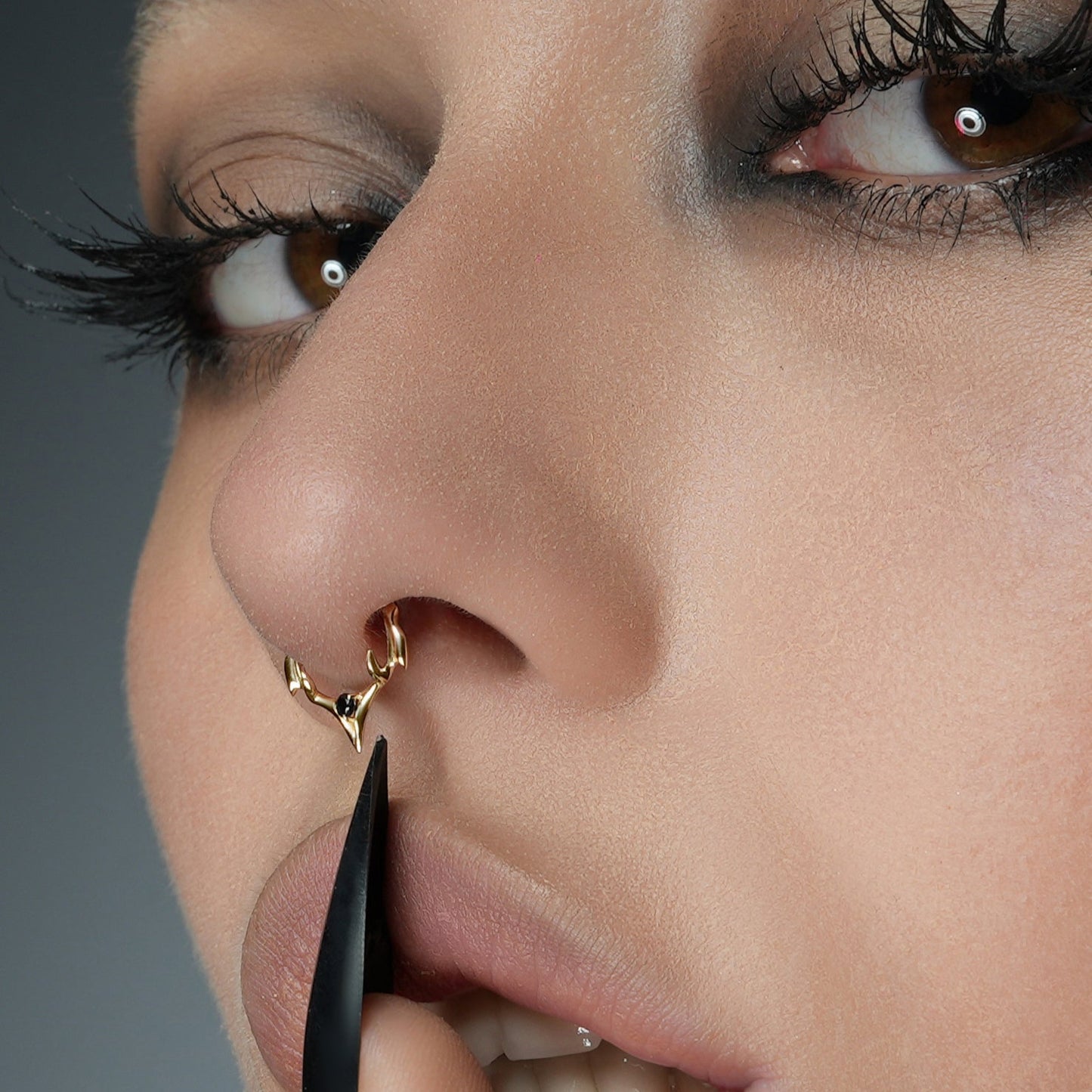 Close-up of a model's nose wearing a gold-tone spiked septum ring with a black gemstone, tweezers adjusting it