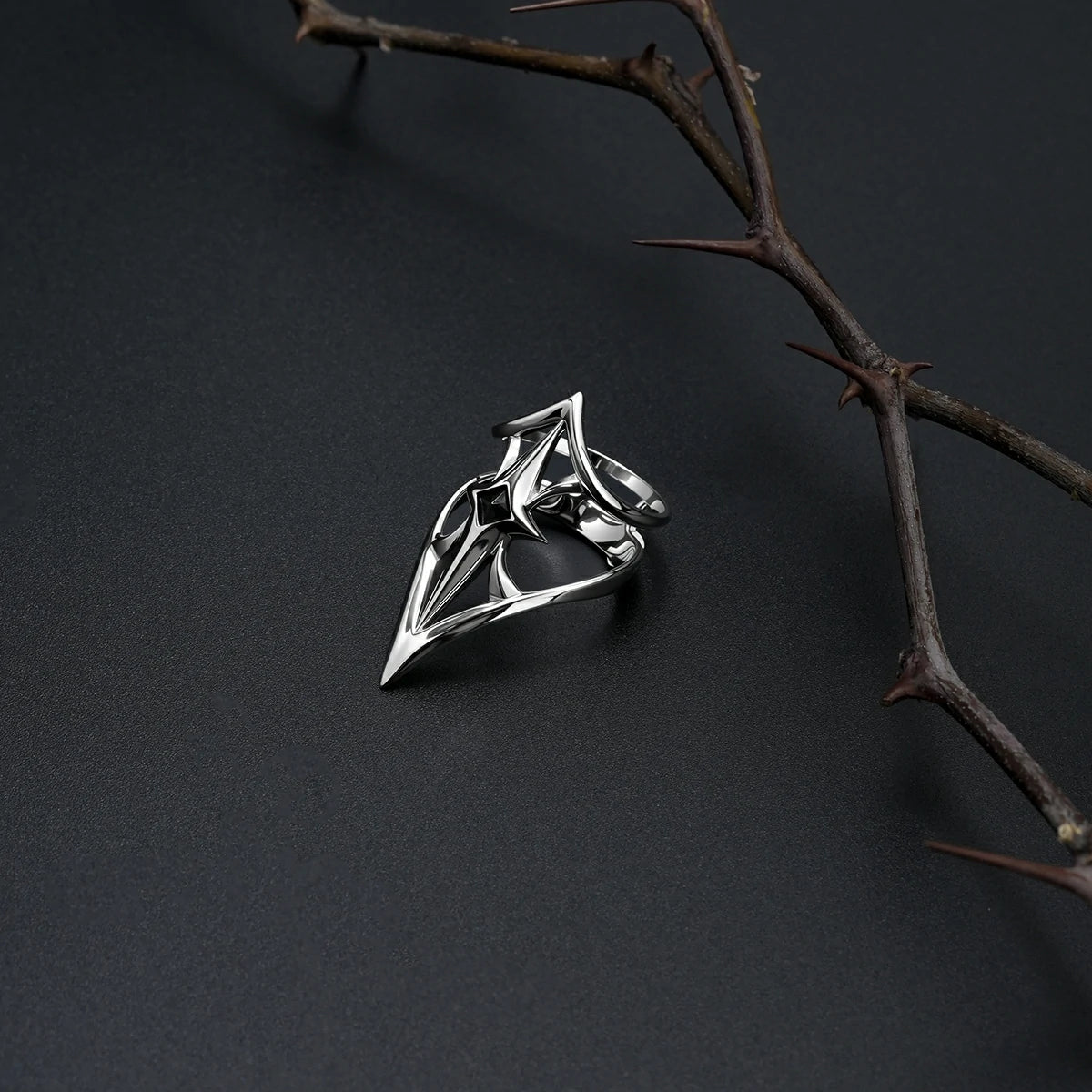Silver-tone Crest Shield ring resting beside a thorn branch on a matte black surface