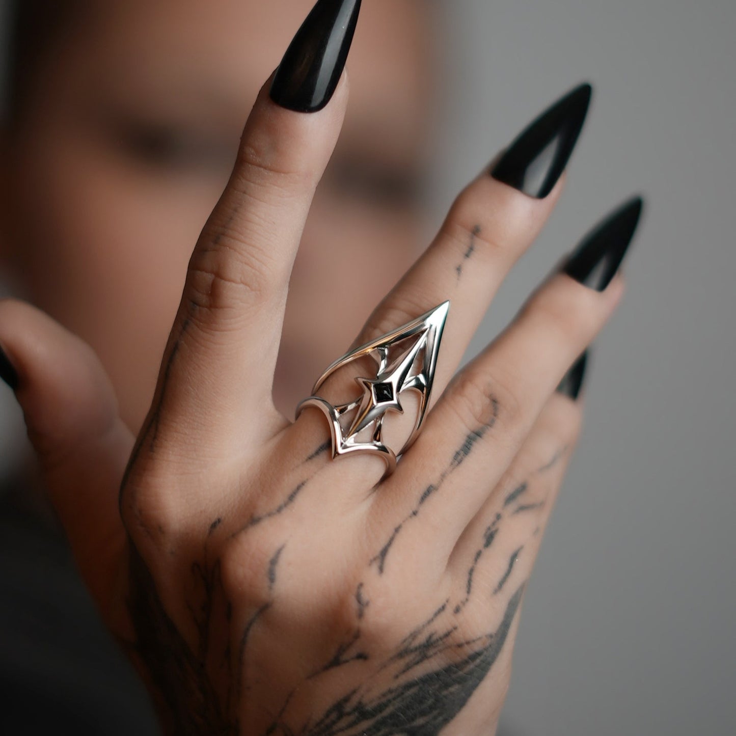 Silver-tone crest shield ring worn on tattooed hand with black stiletto nails, close-up side view