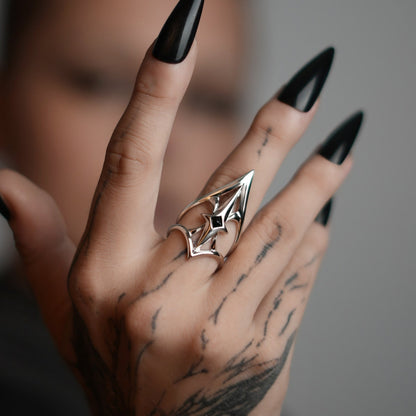Silver-tone crest shield ring worn on tattooed hand with black stiletto nails, close-up side view