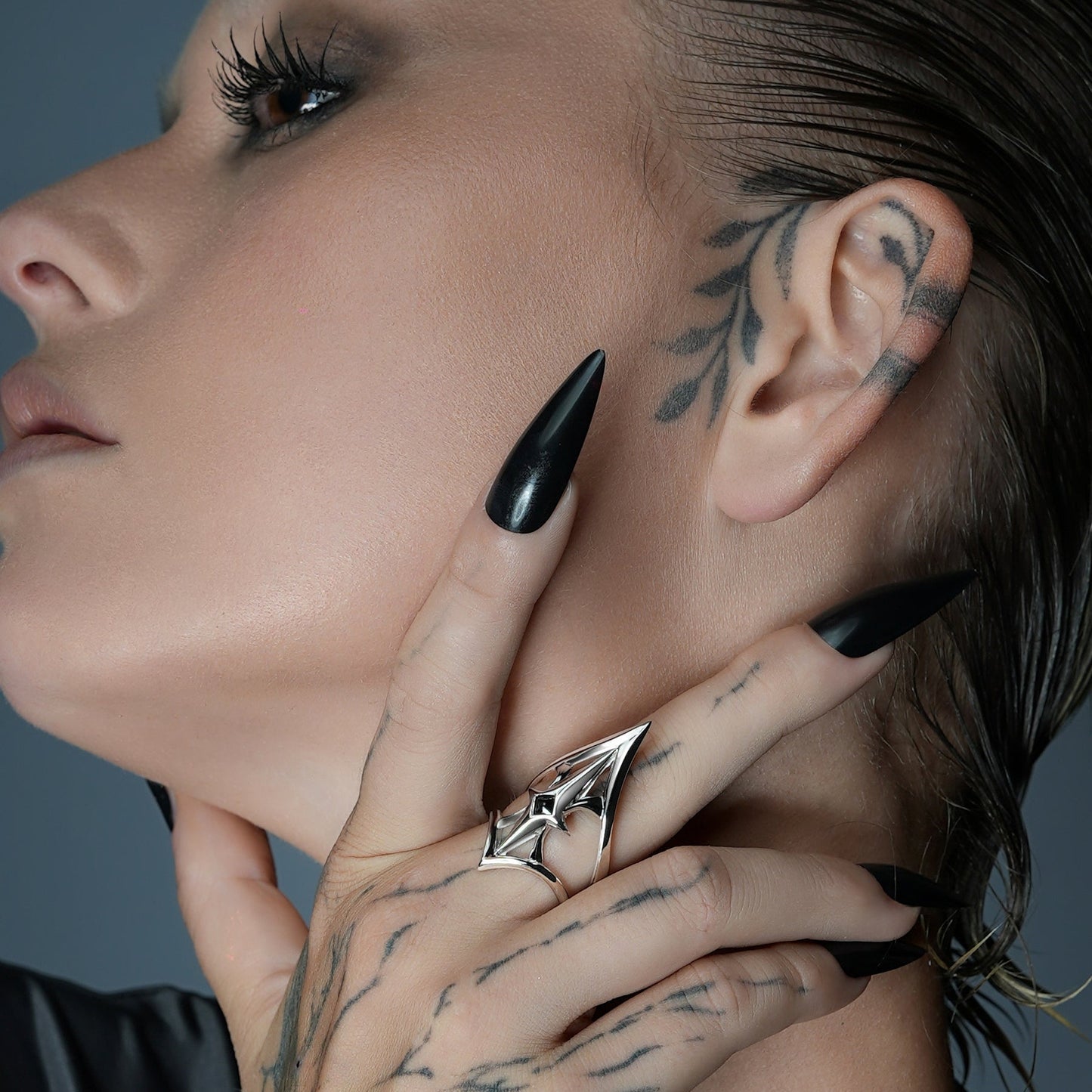 Silver-tone crest shield ring on tattooed fingers resting against a model's neck, profile view