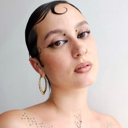 Model wearing a gold-tone marquise hoop earring with engraved patterning alongside a nose ring