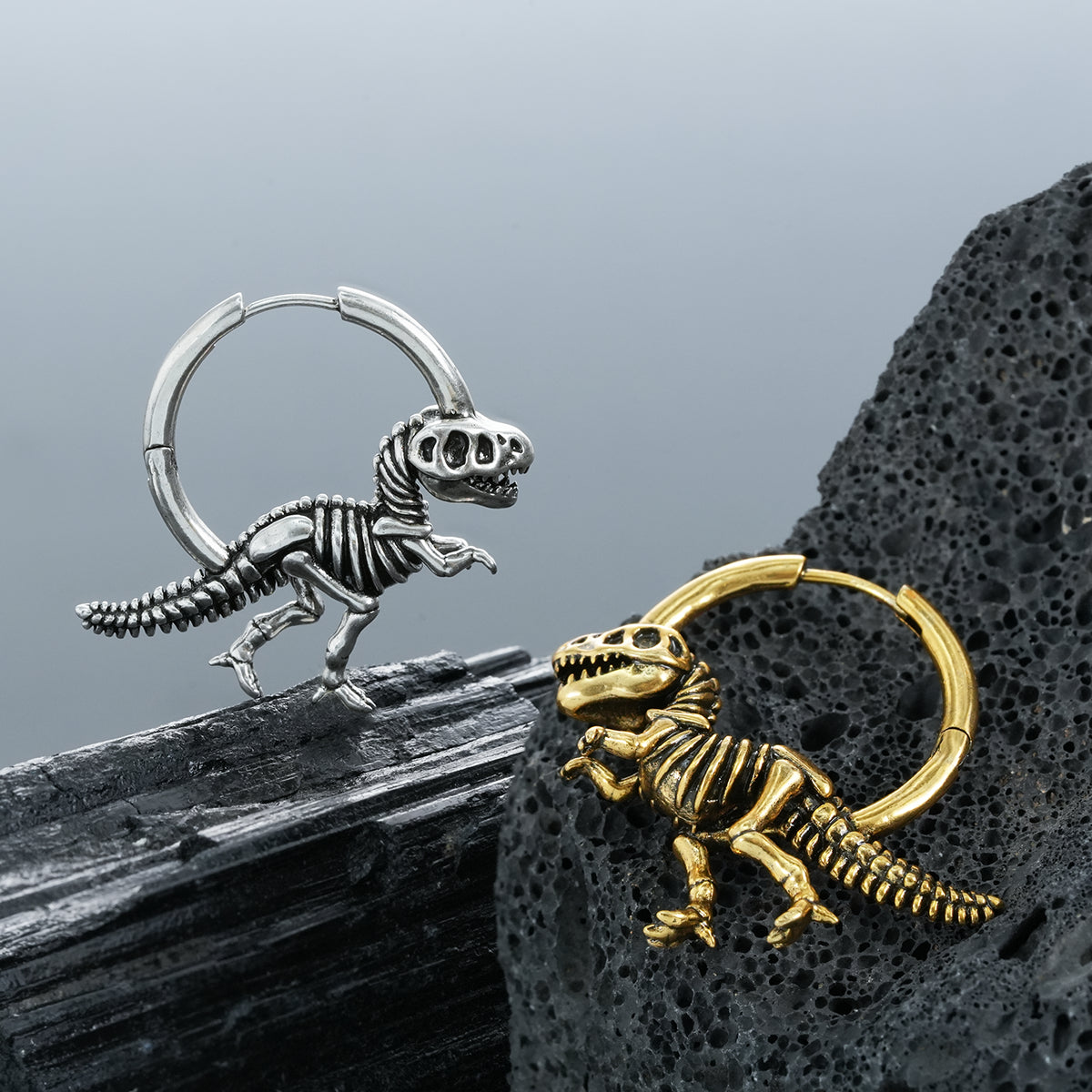 Silver-tone and gold-tone dinosaur skeleton hoop earrings resting on textured black stone surfaces