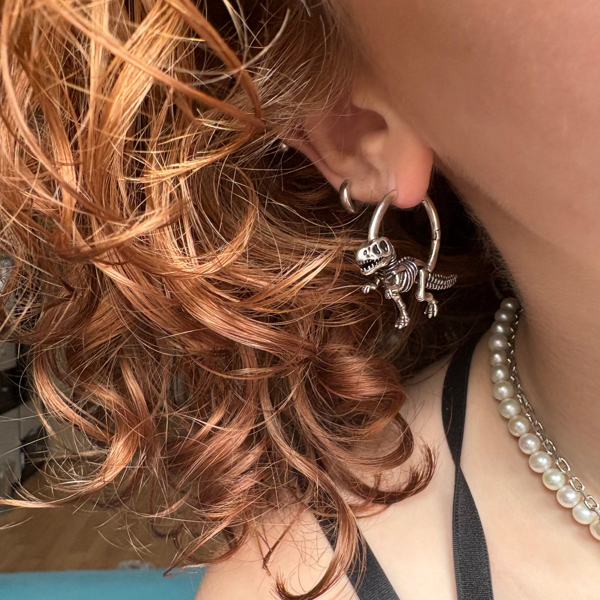Close-up of a model wearing a silver-tone hoop earring with a dinosaur skeleton charm, styled with pearl necklace
