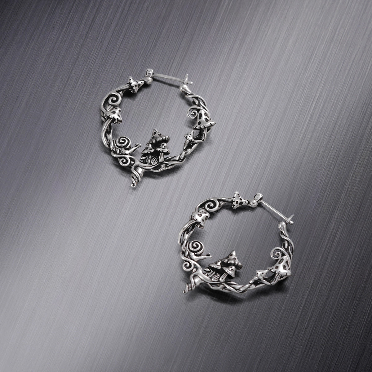 Two silver-tone hoop earrings with mushroom, snail, and vine details, laid flat on a brushed metal surface