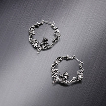 Two silver-tone hoop earrings with mushroom, snail, and vine details, laid flat on a brushed metal surface