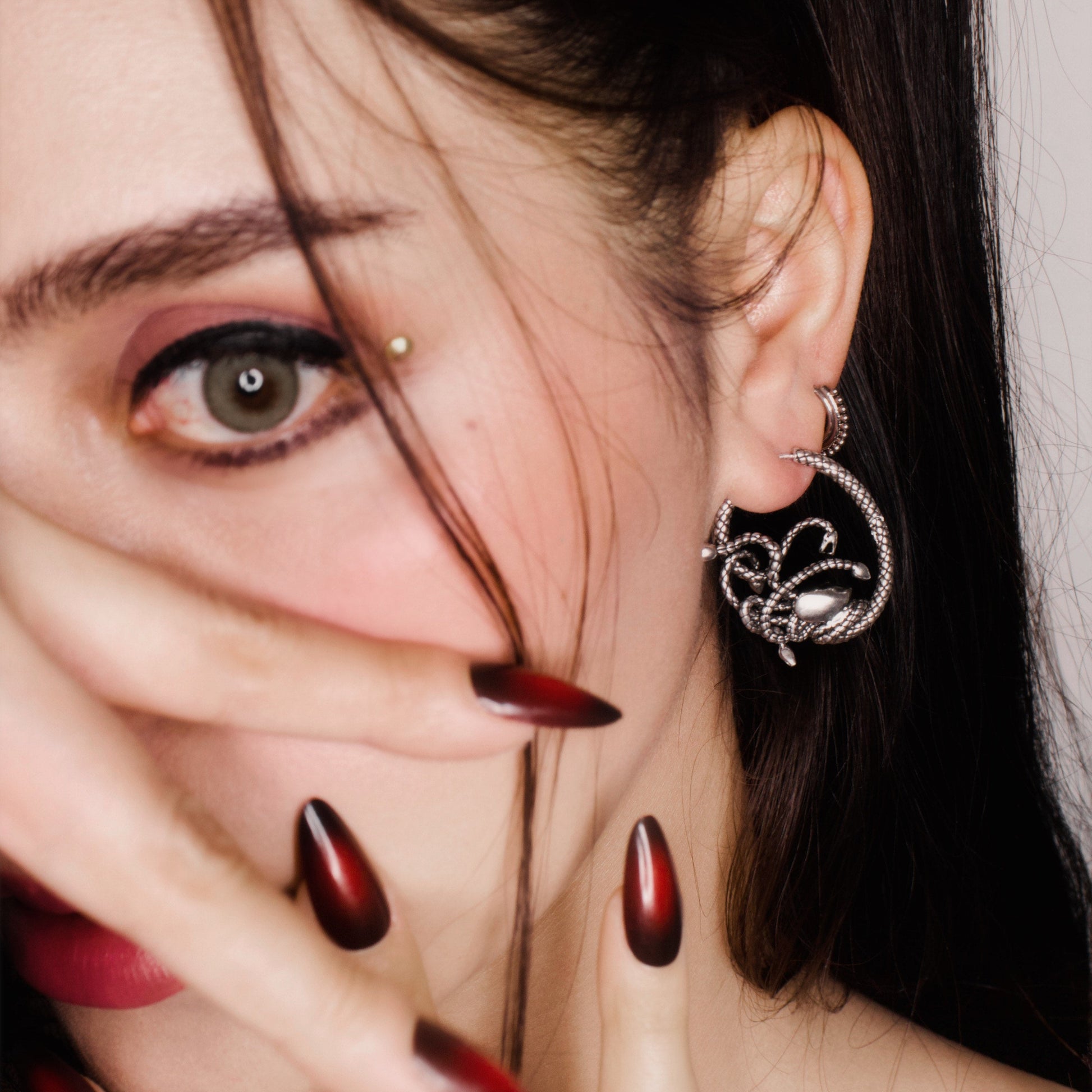Model with dark hair wearing a silver-tone snake motif hoop earring with a tiny face charm, close-up profile view