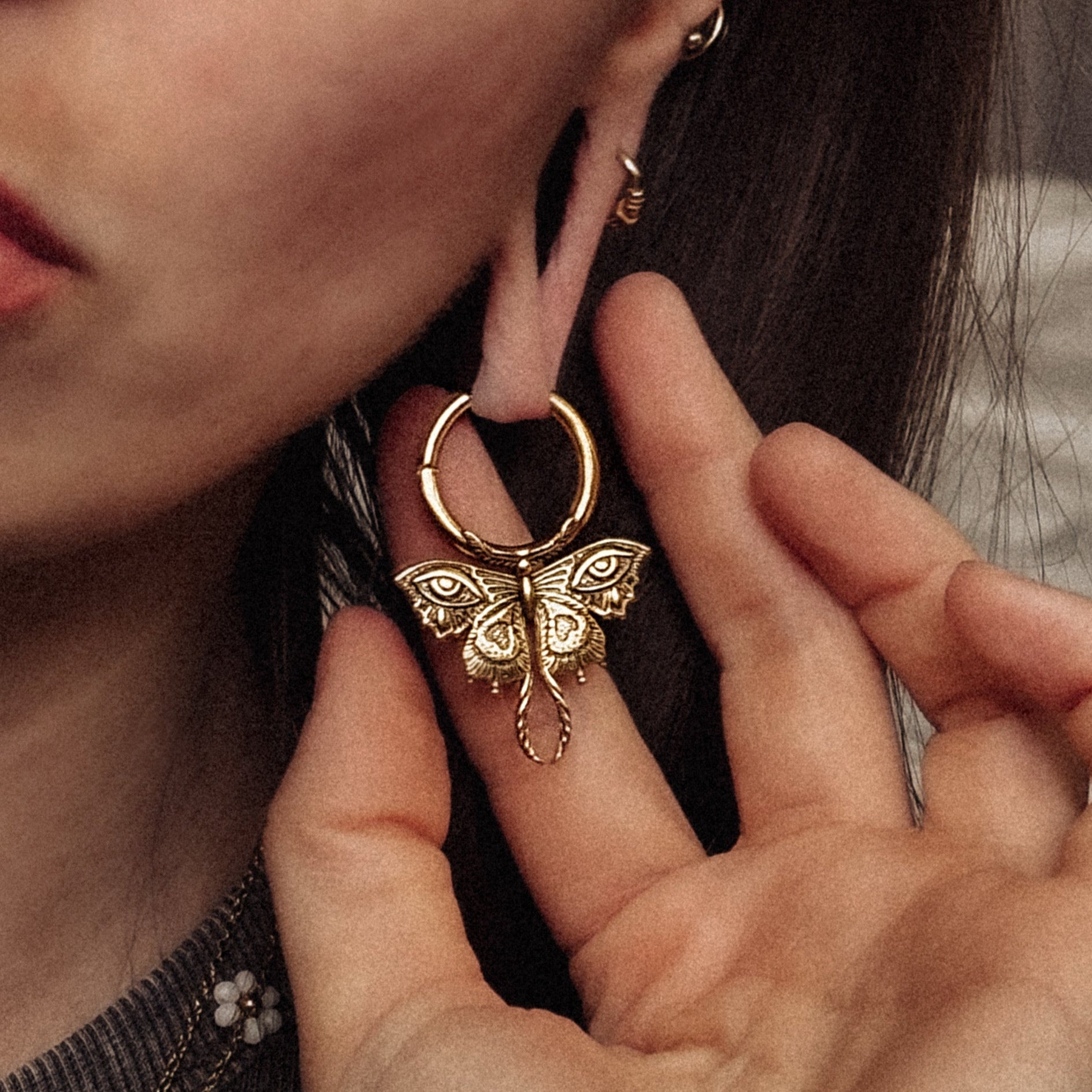 Gold-tone Enchanted Moth hoop earring held beside a model's ear, showing the engraved moth pendant