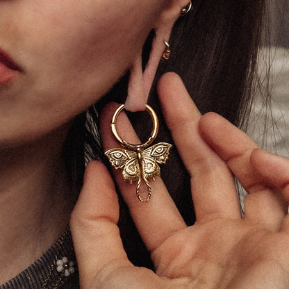 Gold-tone Enchanted Moth hoop earring held beside a model's ear, showing the engraved moth pendant