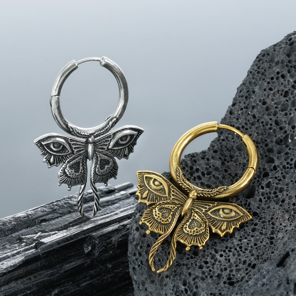 Silver and gold-tone Enchanted Moth hoop earrings displayed on dark volcanic stones