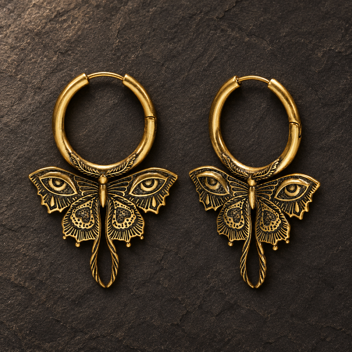 Pair of gold-tone Enchanted Moth hoop earrings with engraved eye wings and curled tails on dark stone.