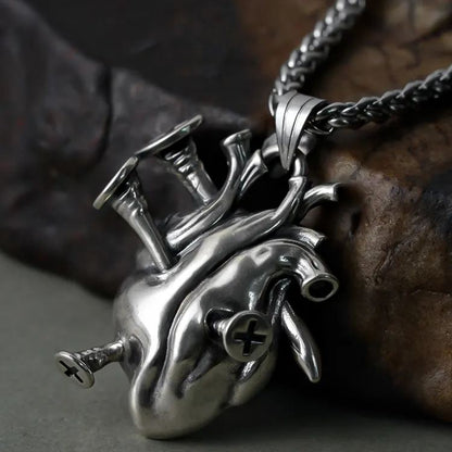 Close-up of a silver-tone anatomical heart pendant with cross screw details on a wheat chain, resting on rock