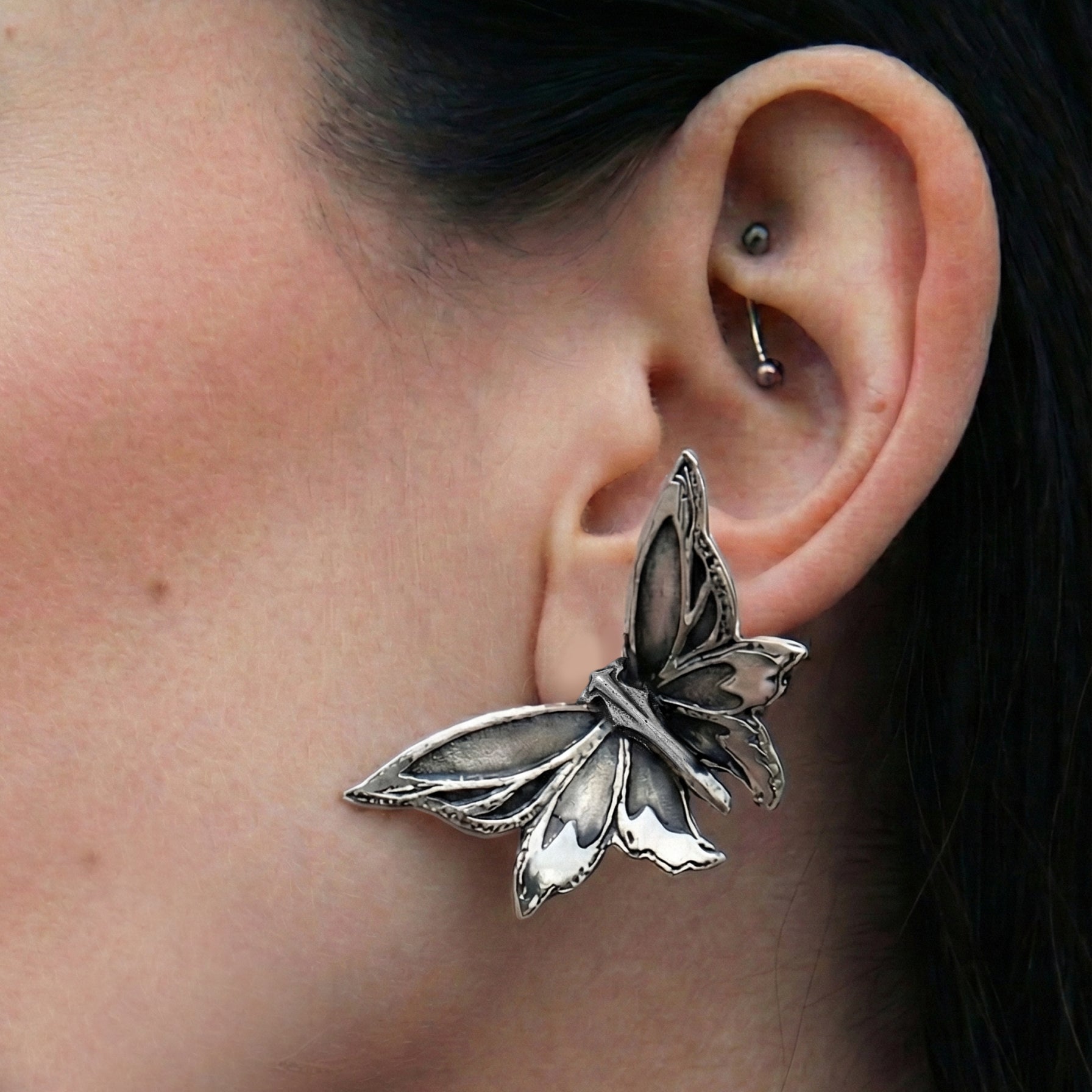 Silver-tone moth ear stud worn on earlobe, close-up side view showing detailed wing texture