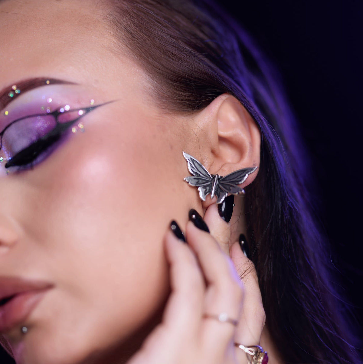 Moth-shaped ear stud in oxidized silver-tone worn on earlobe, close-up under purple lighting