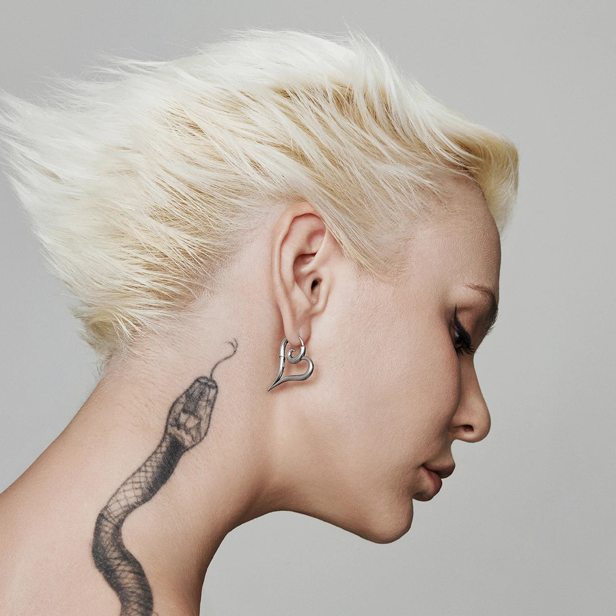 Silver-tone heart hoop earring worn on a model with windswept blonde hair and a snake tattoo on the neck