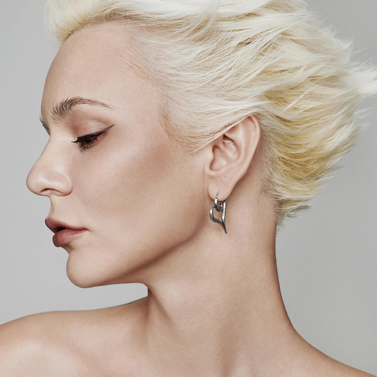 Side profile of a model wearing a silver-tone heart-shaped hoop earring with short platinum hair