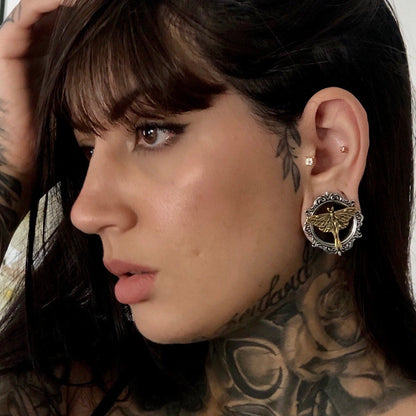 Close profile of tattooed model showing a filigree-edged ear tunnel with gold-tone moth motif on the lobe