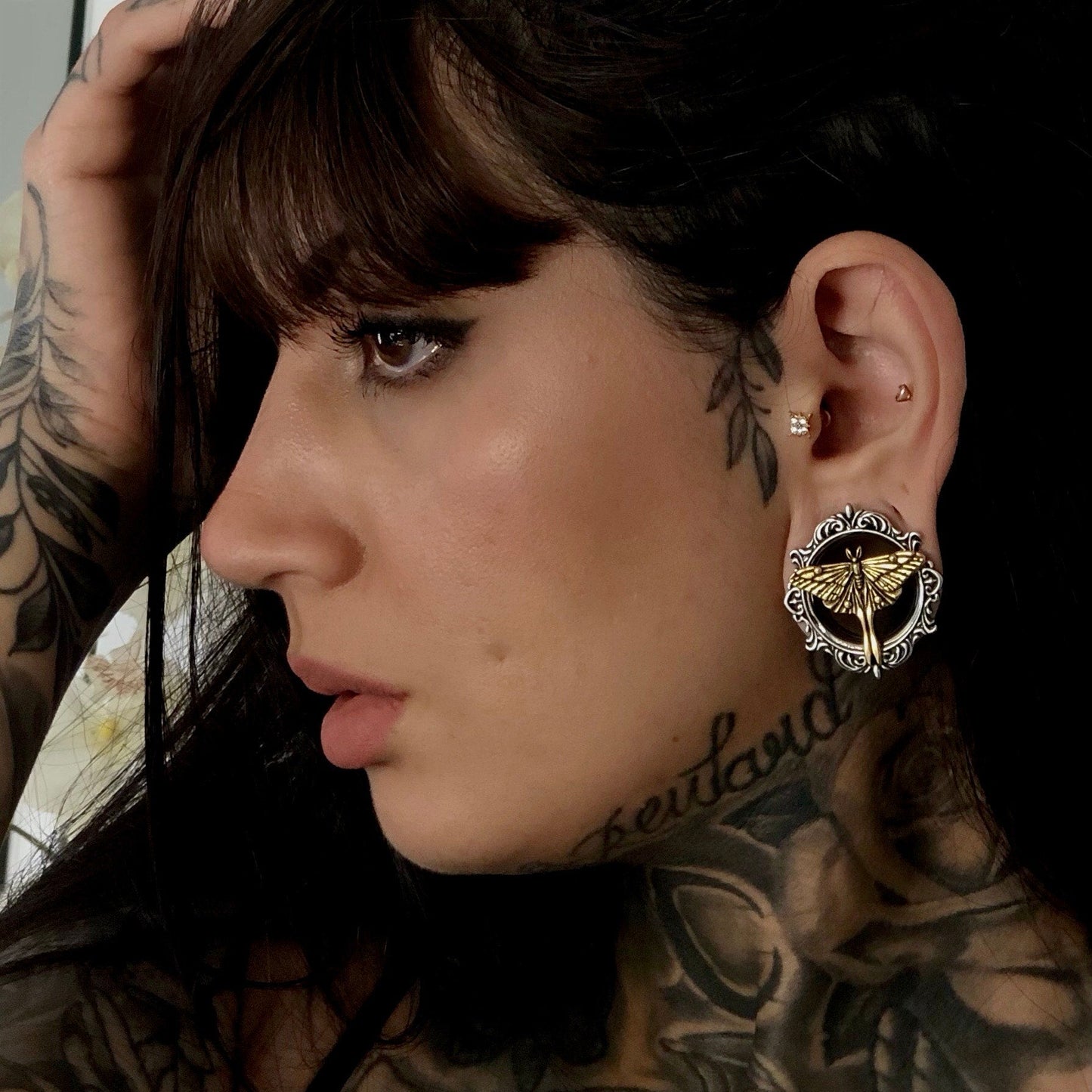 Tattooed model wears an ornate silver-tone ear tunnel with gold-tone moth design in lobe, profile view