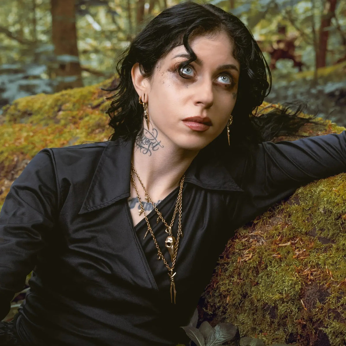 Eye Dagger Earrings in 925 sterling silver, worn in a woodland portrait with dark clothing, layered necklaces, and mossy tree