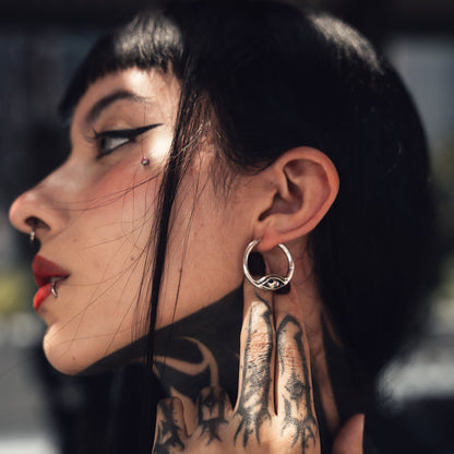 Silver hoop earring with an eye motif worn on a model's ear, side profile view with tattooed hand near the face