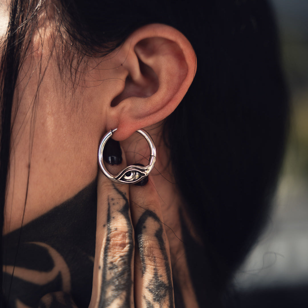 Silver-tone eye motif hoop earring worn on a model, shown against tattooed skin and dark hair