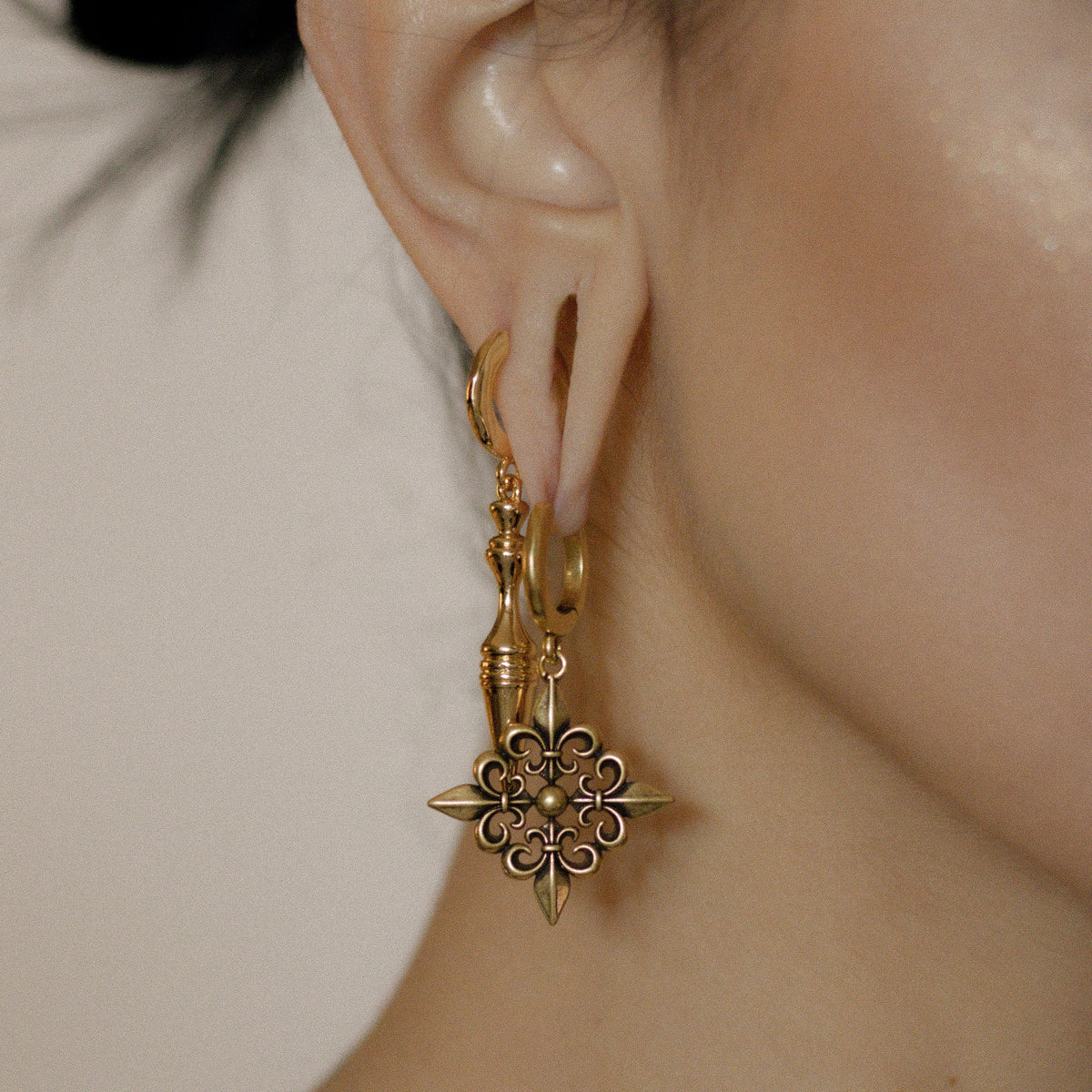 Gold-tone Fleur-de-Lis Elegance hoop earring worn in side profile beside a model's raised hand
